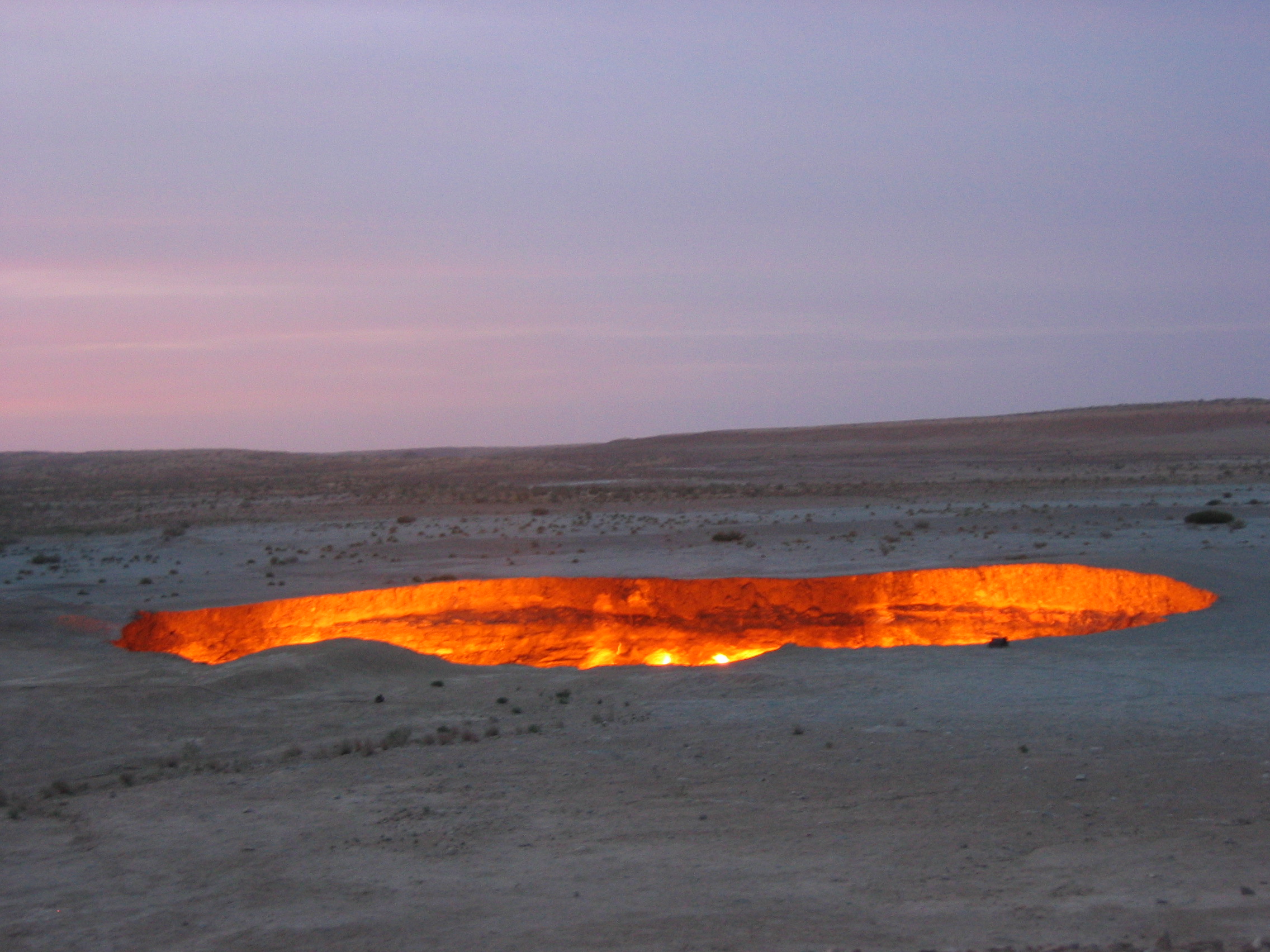 Large glowing crater burning with orange fire in a flat desert landscape at dusk under a pink and purple sky