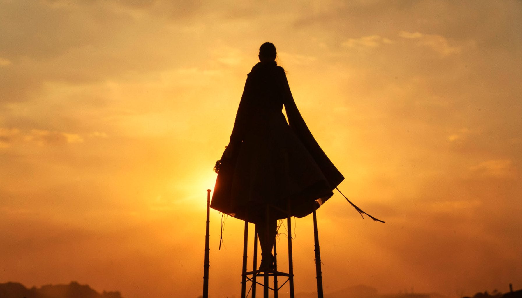 Silhouetted figure on towering stilts against a golden sunset sky, long flowing costume, dramatic and mysterious mood