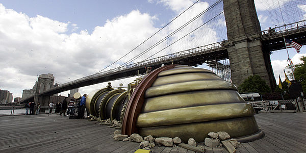 Large curved metallic sculpture on a waterfront boardwalk near a suspension bridge, cloudy sky, industrial mood
