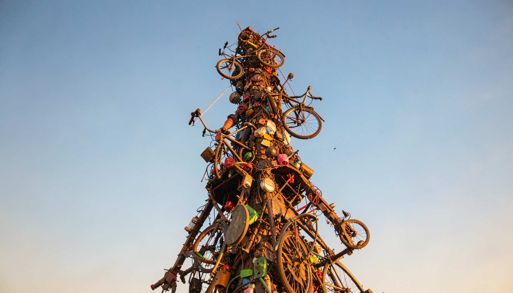 A towering sculpture made of welded scrap metal, bicycle parts, and colorful found objects rising against a bright blue sky